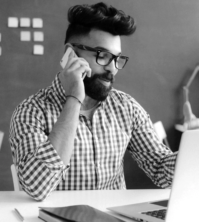 A man with a beard and glasses talking on a phone at a desk